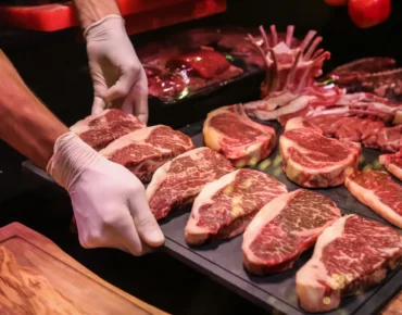 Mano con guantes sosteniendo un plato con cortes de carne roja de res, exhibiendo su veteado y frescura, en un entorno de carnicería.